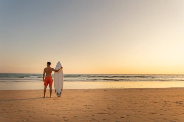 Gün batımında elinde sörf tahtasıyla kıyıda duran atletik vücutlu bir adam. Arkadan bakıyor. Orta çekim