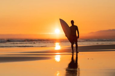 Sahilde elinde turuncu gün batımında sörf tahtası tutan çekici bir adamın siyah silueti. Orta çekim