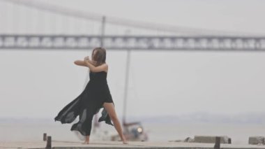 Siyah elbiseli bir kadın yavaş çekimde iskelede dans ediyor. Arka planda bir tekne geçiyor. Orta çekim