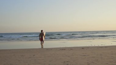 Güneş batarken elinde sörf tahtasıyla okyanusa doğru yürüyen güzel bir atletik vücudu olan bir adam. Orta çekim