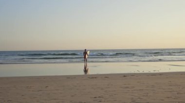 Güneş batarken elinde sörf tahtasıyla okyanusa doğru yürüyen atletik vücutlu bir adam. Orta çekim