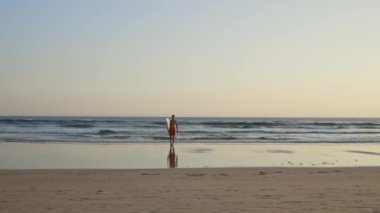 Güneş batarken elinde sörf tahtasıyla okyanustan yürüyen güzel bir atletik vücudu olan bir adam. Orta çekim