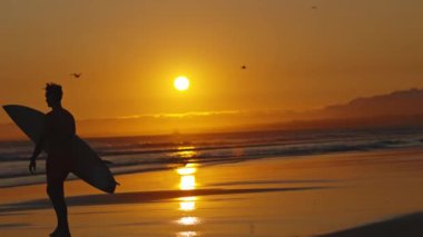 Parlak turuncu gün batımında okyanusa giden bir sörfçünün silueti. Orta çekim