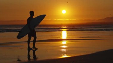 Parlak turuncu gün batımında okyanus kenarında yürüyen bir sörfçünün silüeti. Orta çekim