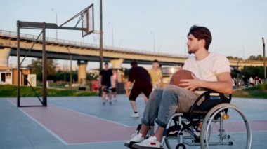 Arka planda basketbol takımı oynarken tekerlekli sandalyedeki bir adam basketbol topunu tutuyor. Orta çekim
