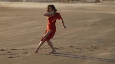 Rüzgarda kumlu sahilde dans eden kırmızı elbiseli zarif genç bayan. Orta çekim