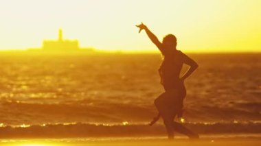 Parlak turuncu günbatımında deniz kıyısında dans eden zarif, güler yüzlü genç bayan. Orta çekim