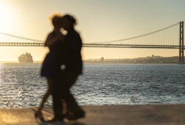 Kucaklaşan ve deniz kenarında dans eden çift silueti. Arka planda büyük bir yolcu gemisi, gün doğumu.