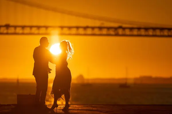 A couples silhouette stands out against a brilliant sunset, with the architectural marvel of a bridge behind.