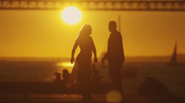 Siluetli çift bir anı paylaşıyor. Gün batımında ya da gün doğumunda dans ediyorlar ve sarılıyorlar. Batan güneş ve ikonik köprü ortamı hazırlıyor..