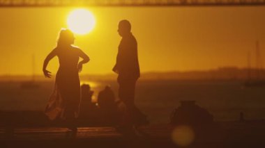 Bir çift silueti, arkasında bir köprünün mimari harikası ile parlak bir günbatımına karşı duruyor..