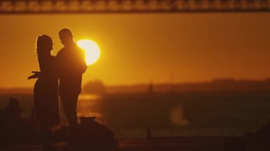 Siluet - bir çift arasında samimi bir kucaklaşma yakalar, gün batımının yumuşak renkleri ve arkaplanda köprü.
