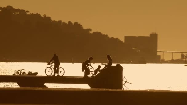 人々のシルエットは公園や川の橋の近くの桟橋に休憩しています ポルトガル アルマダの川の近くのバイサイクルと家族公園の人 黄金の時間 — ストック動画