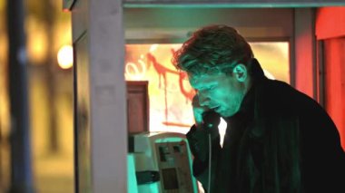 Bir adam telefon kulübesinde konuşuyor. Kabin loş bir alanda.