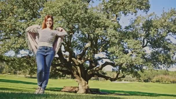 女性が大きな木の前に立って ストライプのシャツとジーンズを着ています シーンは平和で穏やかで 木の前で写真を撮る女性と — ストック動画