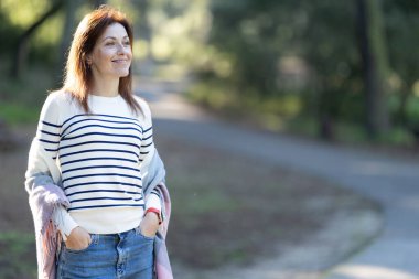 Diş teli takan, çizgili kazak giyen ve kot pantolon giyen yetişkin bir kadın yolda duruyor. Gülümsüyor ve sağına bakıyor.