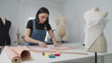 Bir kadın masanın üzerinde elbise tasarımı üzerinde çalışıyor. Masa kumaş, makas ve diğer dikiş aletleriyle kaplı. Kadın işine odaklanmış durumda ve sahne yaratıcılık duygusunu aktarıyor.