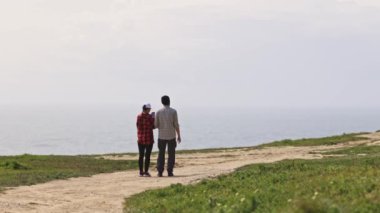 Bir çift okyanus kenarında bir yolda yürüyor. Adam şapka takıyor ve kadın kırmızı bir gömlek giyiyor. Sahne huzurlu ve romantik.