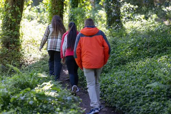 A group of friends, walking in a line, are making their way down a dirt path surrounded by tall trees and green foliage in a wooded area.