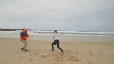 İki kişi plajda sopa savaşı oynuyor. Adam turuncu bir ceket giyiyor ve kadın beyaz bir gömlek giyiyor.