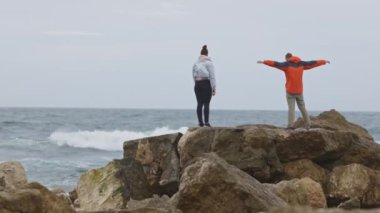 Okyanusa bakan bir kayanın üzerinde duran iki kişi. Bir tanesi kırmızı ceket giyiyor.
