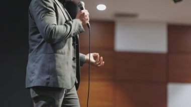 Bir adam elinde mikrofonla sahnede dikiliyor. O bir takım elbise ve kravat giydi ve bir konuşma yapıyor. Profesyonellik ve resmiyet kavramı