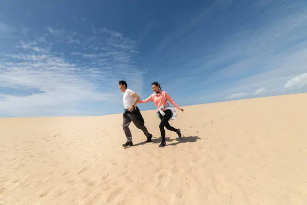 A man and woman are running on a sandy beach. The man is wearing a white shirt and the woman is wearing a pink shirt. The sky is blue and there are some clouds in the background