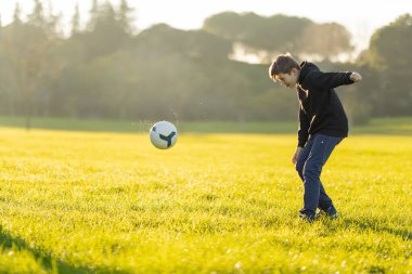 Bir çocuk tarlada futbol topunu tekmeliyor. Oğlan siyah kapüşonlu ve kot pantolon giyiyor.