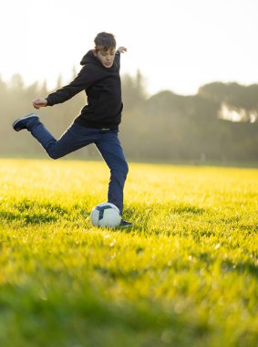 Bir çocuk tarlada futbol topunu tekmeliyor. Oğlan siyah kapüşonlu ve kot pantolon giyiyor.