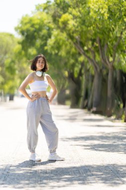 Bir kadın kaldırımda duruyor, kulaklık takıyor ve beyaz bir atlet giyiyor. Sağına bakıyor.