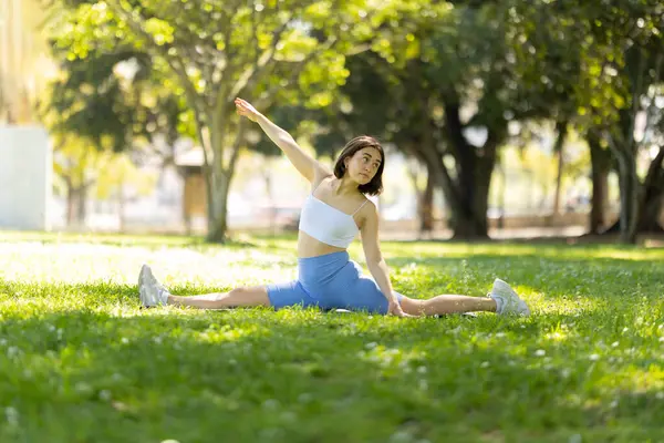 Bir kadın parkta yoga pozu veriyor. O bölünmüş bir pozisyonda ve yukarı bakıyor. Sahne huzurlu ve sakin. Kadınlar duruşuna ve çevreleyen yeşilliğe odaklanıyor.