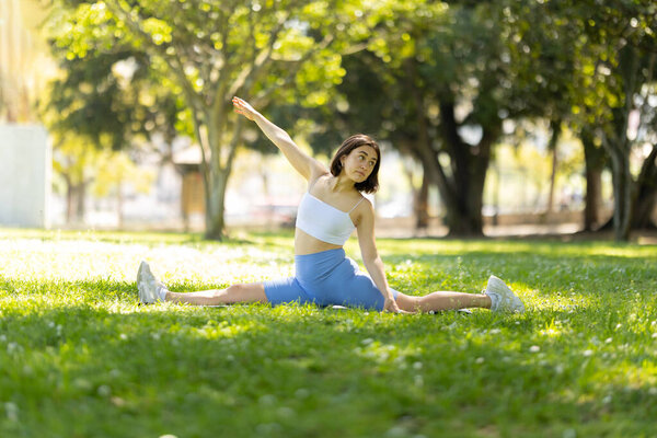 A woman is doing a yoga pose in a park. She is in a split position and is looking up. The scene is peaceful and serene, with the womans focus on her pose and the surrounding greenery