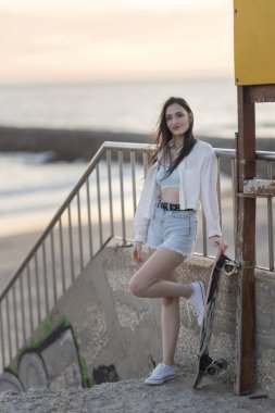 Bir kadın elinde kaykayıyla beton bir çıkıntının üzerinde duruyor. O, beyaz bir gömlek ve mavi bir şort giyiyor. Görüntüde rahat ve rahat bir hava var.