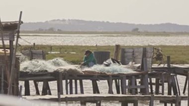 26 Nisan 2024 - Portekiz, Porto Palacio da Carrasqueira - bir kadın iskelede bir balık ağını tamir ediyor. Su sakin ve gökyüzü bulutlu. Adam yeşil bir gömlek giyiyor.