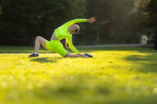ネオングリーンの服を着た女性は 草原でヨガのポーズをしています 彼女の衣装と緑の草の明るい色は 陽気でエネルギッシュな気分を作り出しています 健康と健康に関するコンセプト — ストック写真