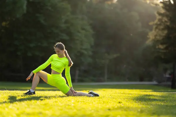 一个穿着霓虹灯黄色衣服的快乐女人正在公园的草地上悠闲地伸展着双腿 四周环绕着郁郁葱葱的绿叶和其它享受大自然的人们 — 图库照片
