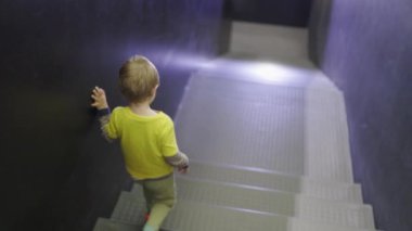 Küçük bir çocuk merdivenleri çıkıyor. Çocuk sarı bir gömlek giyiyor ve duvara tutunuyor. Çocuk merdivenleri keşfederken macera ve merak kavramı