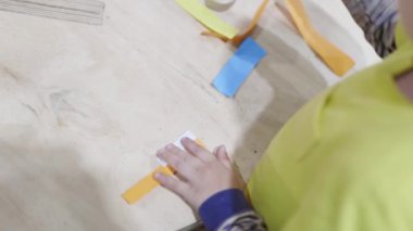 Bir çocuk üzerinde beyaz çıkartma olan bir kağıt şerit yapıyor. Çocuk sarı bir gömlek giyiyor.