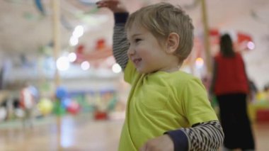 Sarı tişörtlü genç bir çocuk renkli oyuncaklarla dolu bir odada duruyor. Gülümsüyor ve eğleniyor.