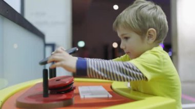 Genç bir çocuk parlak ışıklı bir odada bir oyuncakla oynuyor. Çocuk sarı bir gömlek giyiyor ve oyuncağa odaklanmış. Oda bir klavye de dahil olmak üzere çeşitli oyuncaklar ve nesnelerle dolu.