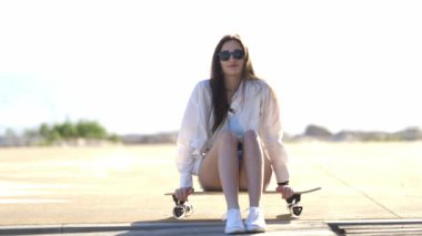 Kaykayıyla yerde oturan bir kadın var. Beyaz bir gömlek ve şort giyiyor.