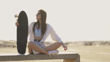 Elinde kaykayla bankta oturan bir kadın var. Beyaz bir gömlek ve şort giyiyor.