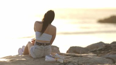 Bir kadın okyanus kıyısındaki bir kayanın üzerinde oturuyor, denize bakıyor. Beyaz bir atlet ve beyaz bir şort giyiyor.