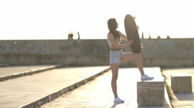 Bir kadın elinde kaykayla bir çıkıntının üzerinde duruyor. Sahne arka planda birkaç kişinin olduğu bir parkta geçiyor. Sahne rahat ve sıradan.