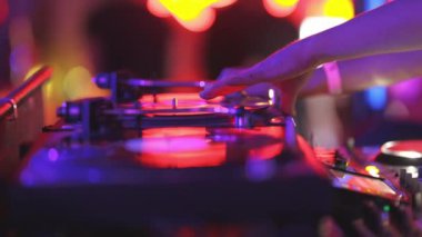 Kırmızı ve mavi arkaplanı olan bir DJ seti çalıyor. Müzik gürültülü ve enerjiktir, ve kişi elindeki işe odaklanır. Atmosfer hareketli ve eğlenceli, insanlar dans ediyor.