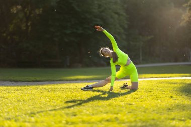 Neon yeşili kıyafetli bir kadın çimenli bir alanda yoga pozu veriyor. Kıyafetinin parlak renkleri ve çevreleyen yeşil çimenler neşeli ve enerjik bir atmosfer yaratıyor.