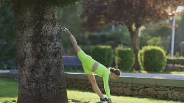 Bir kadın bir ağacın önünde bacağını esnetiyor. Rahatlama ve sükunet kavramı, kadın dışarıda bir parkta, yeşilliklerle çevrili.