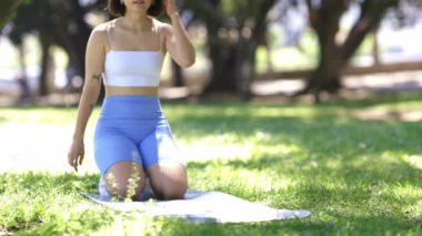 Bir kadın parkta minderde yoga yapıyor. Mavi şort ve kolsuz bluz giyiyor.