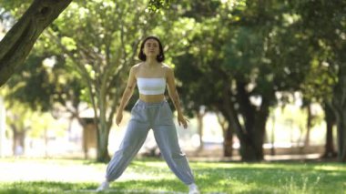 Bir kadın parkta yoga pozu veriyor. Park ağaçlar ve otlarla dolu, huzurlu ve huzurlu bir atmosfer yaratıyor. Kadın vücudunu esnetirken odaklanmış ve kararlı.