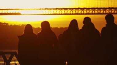 Birkaç kişi bir bankta oturup güneşin batışını izliyor. Arka planda Tagus feribotu geçiyor..
